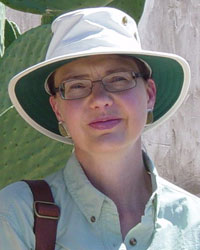 a woman with a bucket cap, glasses, and a tan jacket looks at the camera