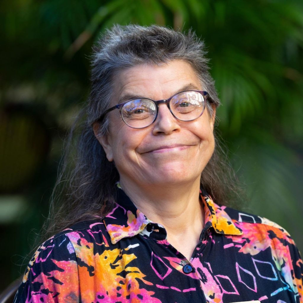 a woman in a colourful shirt with glasses and grey hair smiles