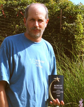 a balding man with a blue shirt and grey facial hair stands in front of a bush