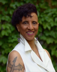 a woman with a white sleeveless jacket, hanging earrings, and styled black hair smiles