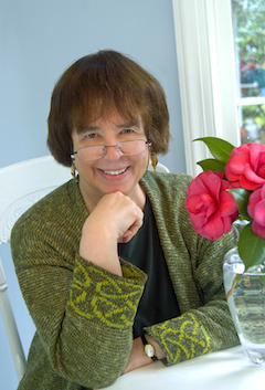 a woman in a green jacket and glasses sits at a table with a vase of red flowers