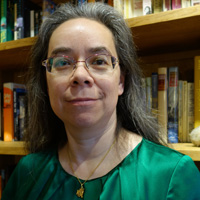 a woman with a green blouse, a pendant necklace, grey hair, and glasses smiles