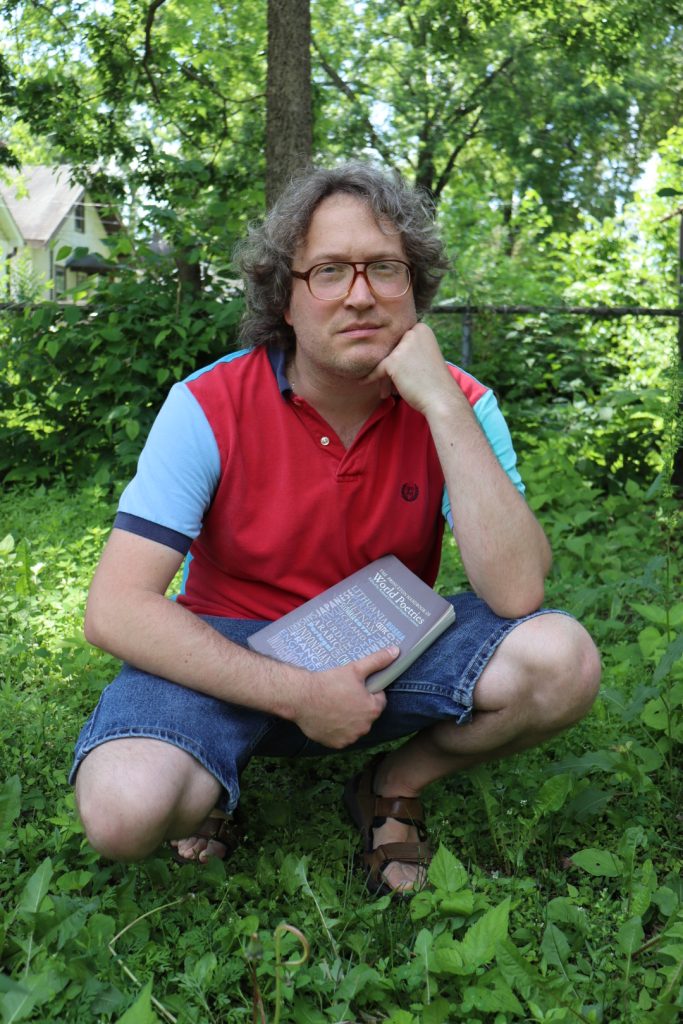 a person in a red coat and glasses crouches in front of greenery, holding a book